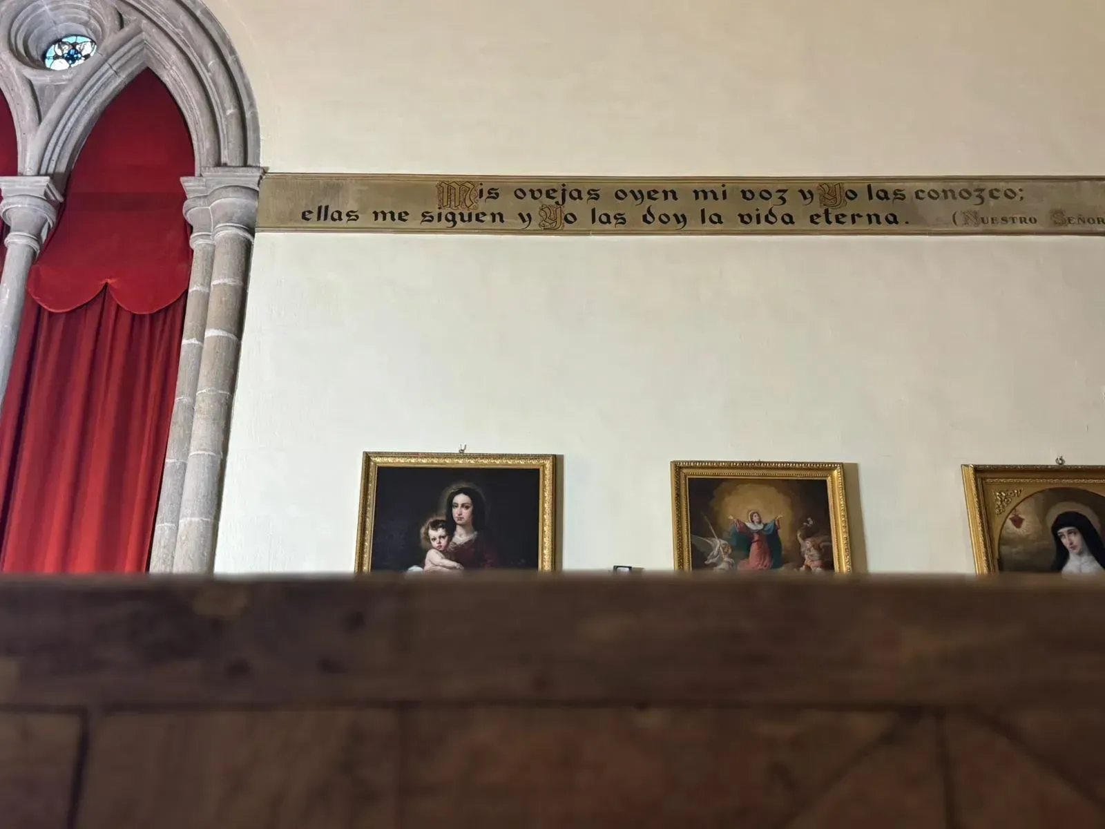 Interior de iglesia con pinturas bajo una inscripción. Cortina roja a la izquierda y ventanas arqueadas.