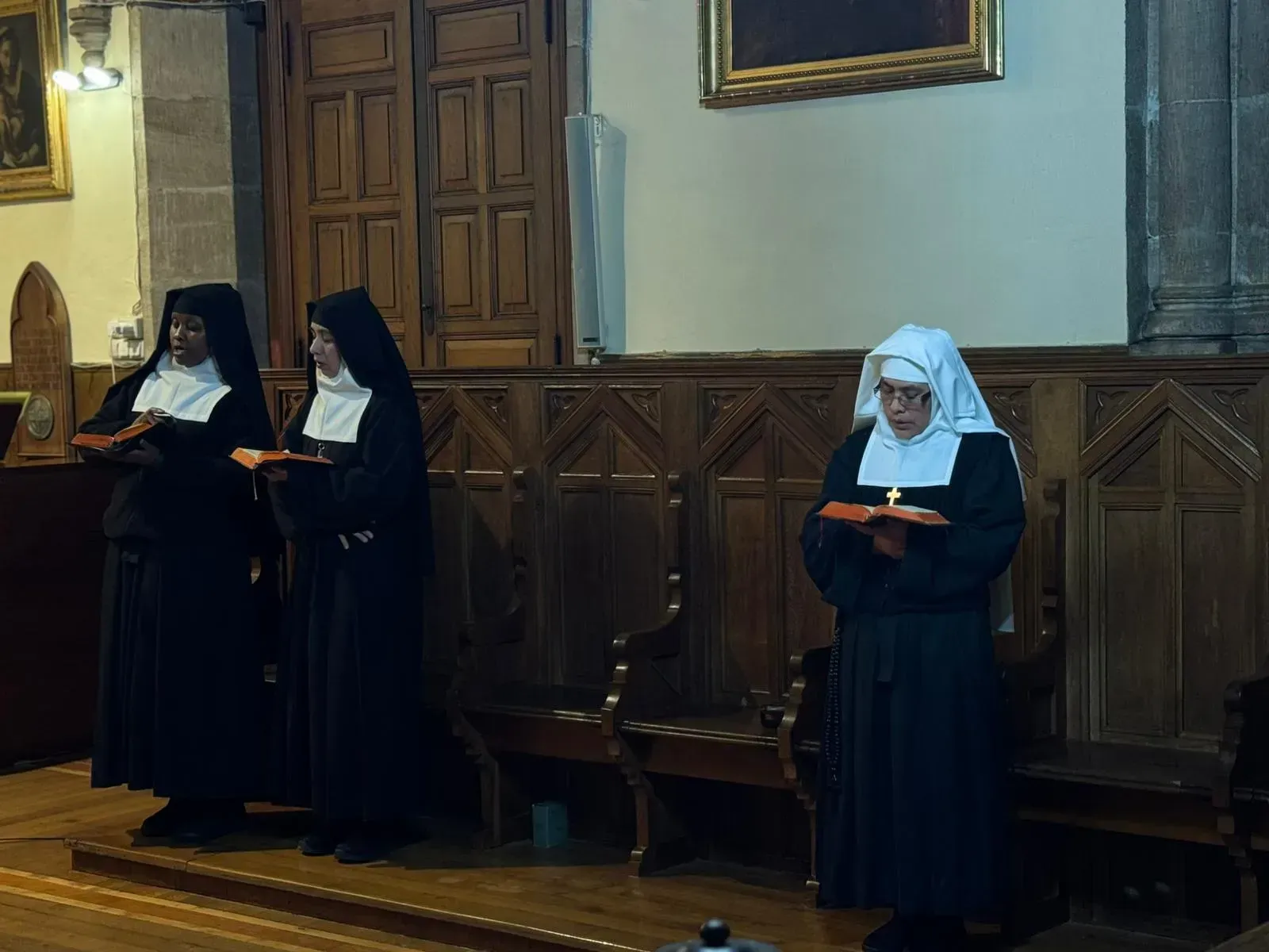 Tres monjas con hábitos negros y velos blancos leen libros en una iglesia.