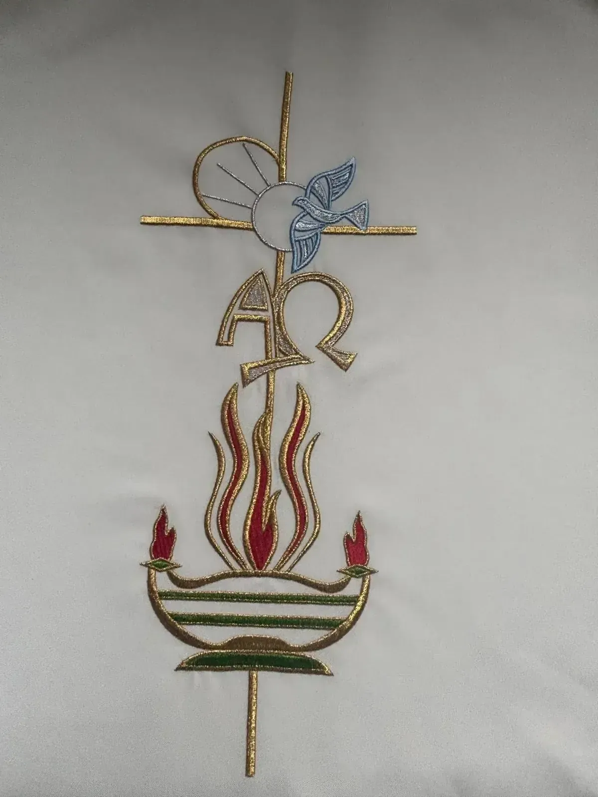 Símbolo religioso bordado sobre tela blanca: cruz, paloma, llama, Alfa y Omega, detalles dorados y azules.