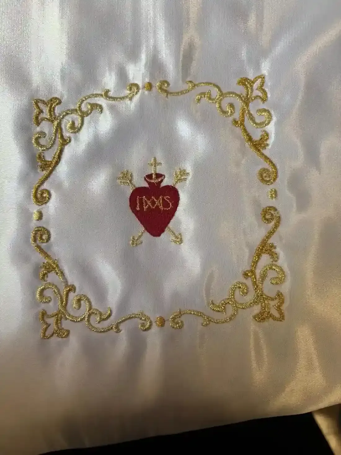 Símbolo del Sagrado Corazón bordado en oro y rojo, rodeado de un borde decorativo dorado sobre tela blanca.