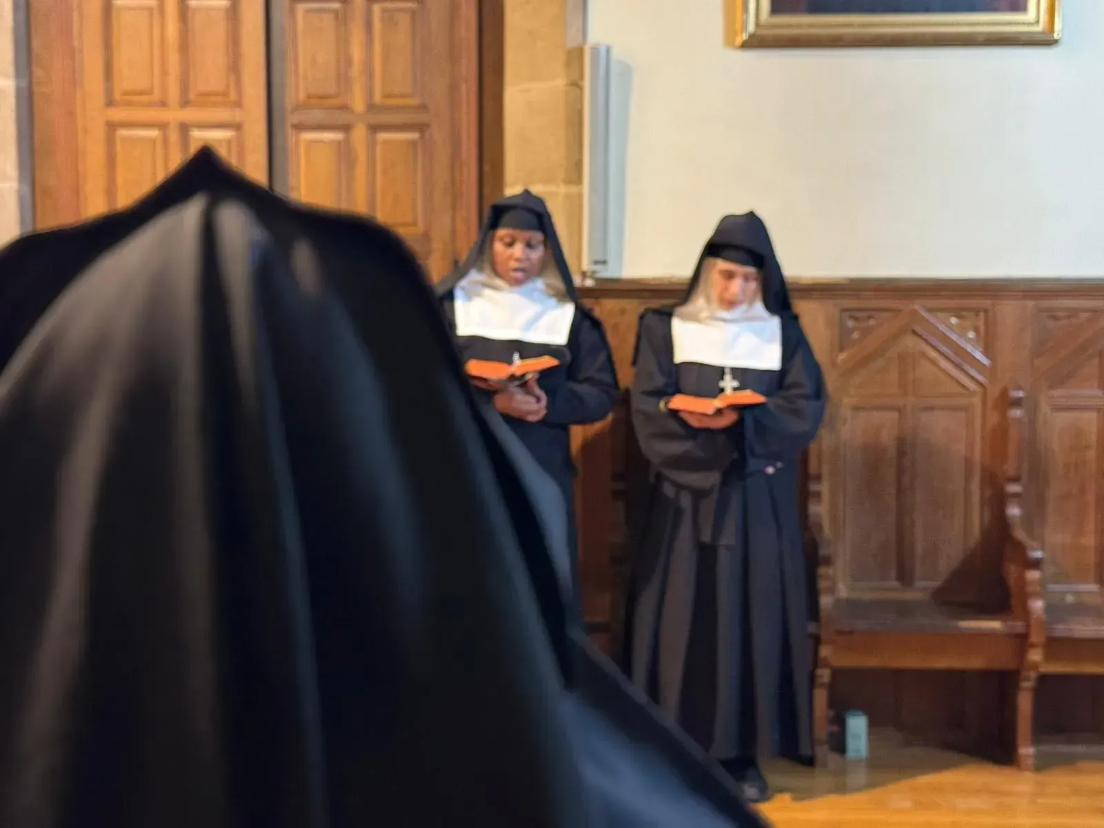 Dos monjas con hábitos negros y cuellos blancos, rezando con libros en una habitación de madera.