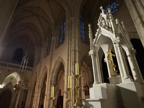 Vista interior de una catedral con arquitectura gótica; arcos, vidrieras y un crucifijo dorado.