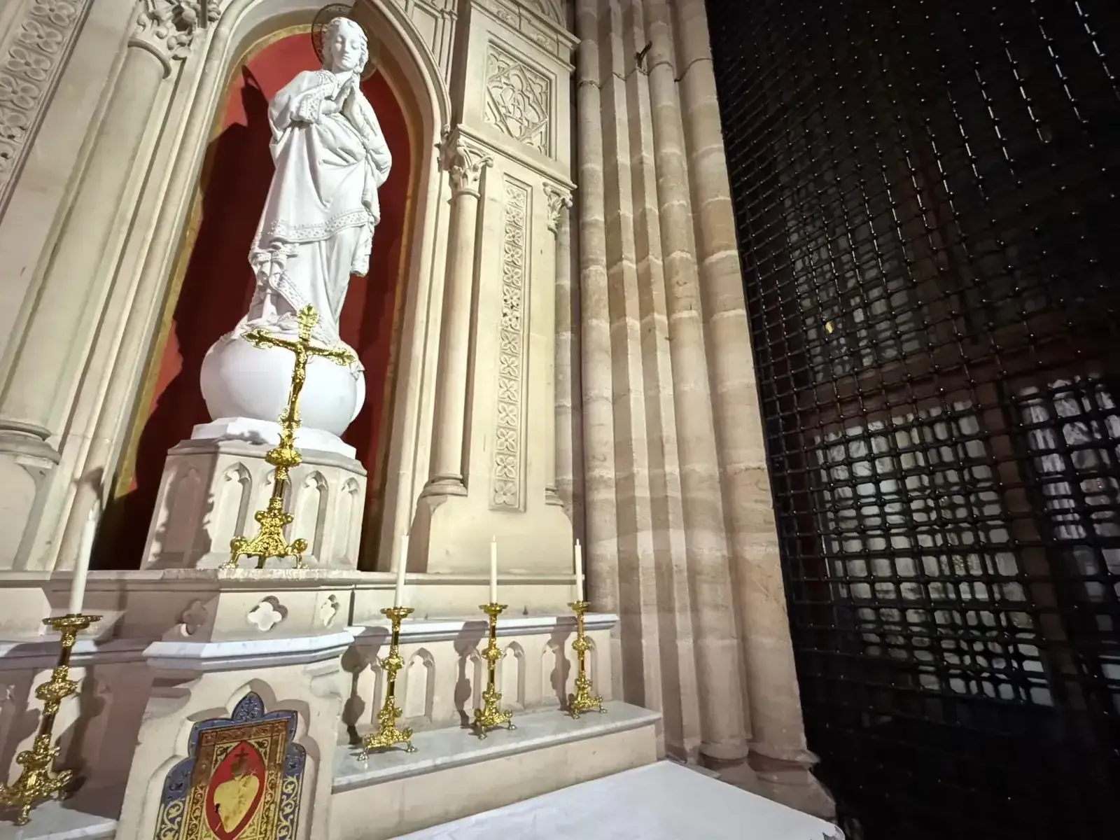 Estatua de un santo sobre un pedestal en una hornacina de una iglesia con una cruz de oro, velas y detalles arquitectónicos góticos.