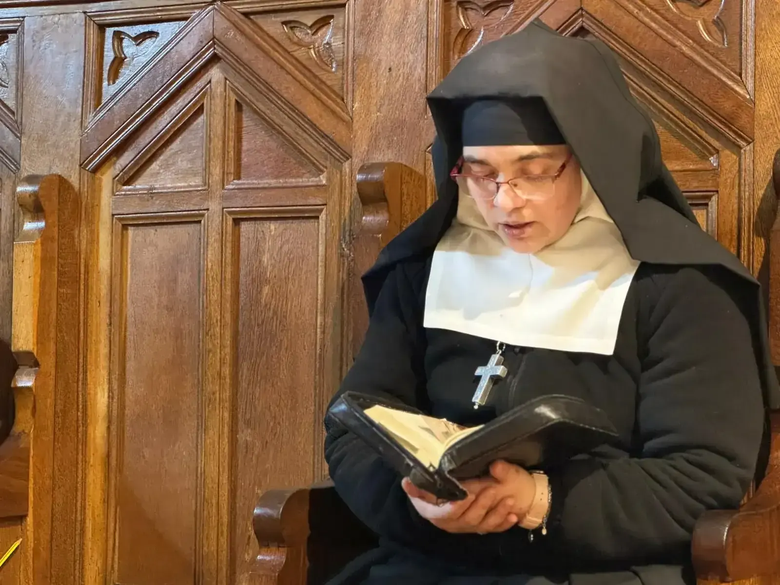 Monja leyendo un libro en un banco de madera de una iglesia, usando gafas y un crucifijo.