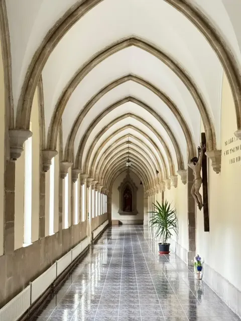 Largo pasillo arqueado en una iglesia o monasterio, con pilares de piedra, ventanas y obras de arte religiosas al final.