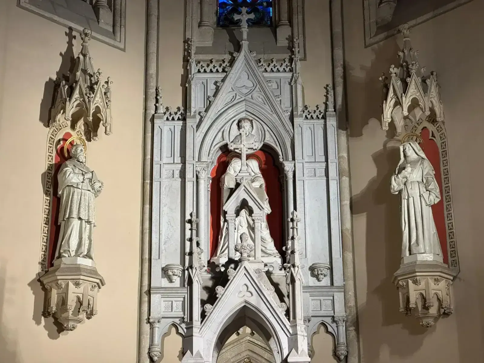 Esculturas en hornacinas de iglesias. La figura central sostiene una cruz, flanqueada por santos. Piedra clara, detalles en rojo y paredes beige.