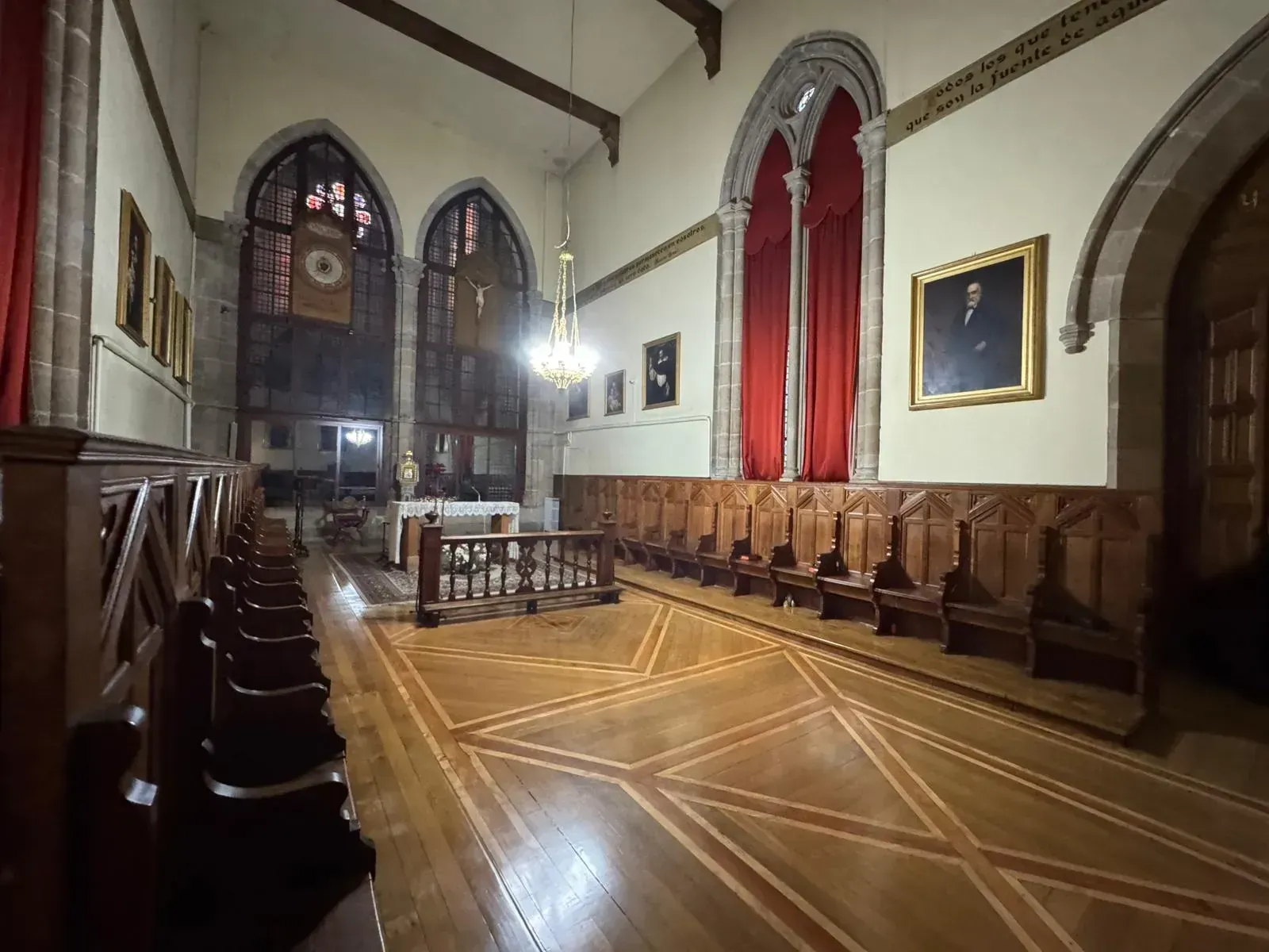 Interior de una capilla con bancos de madera, vitral ornamentado, altar y retratos en las paredes.