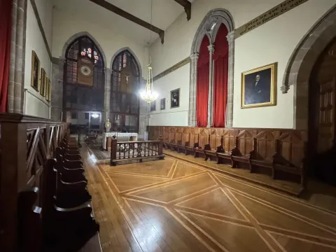 Interior de una capilla con bancos de madera, vitral ornamentado, altar y retratos en las paredes.
