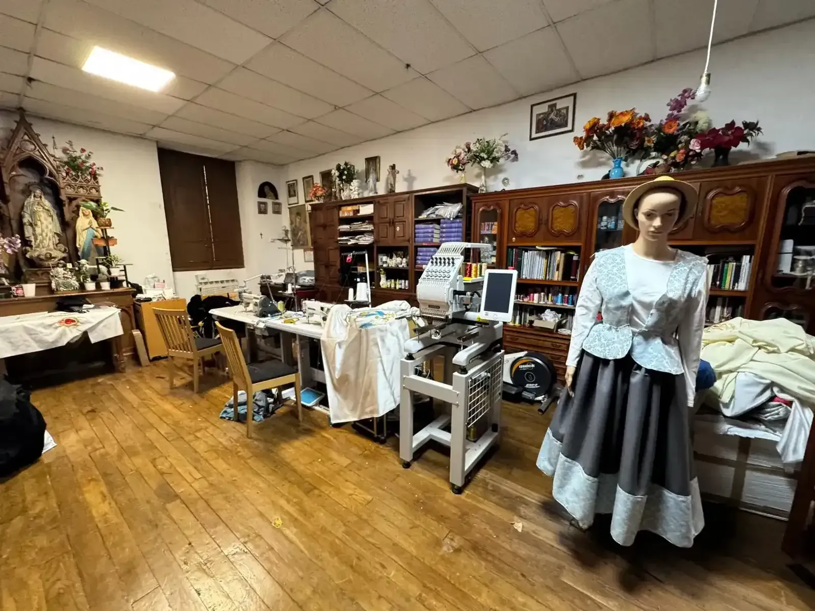 Un maniquí con un vestido se encuentra en un taller desordenado con mesas de costura, armarios y artículos religiosos.