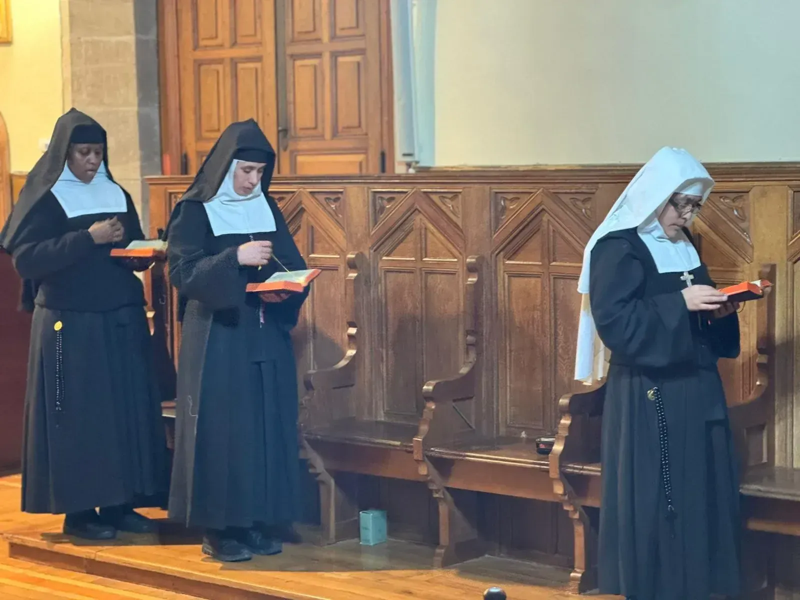 Tres monjas con hábitos negros y velos blancos están de pie, leyendo libros de oraciones en un entorno de iglesia de madera.