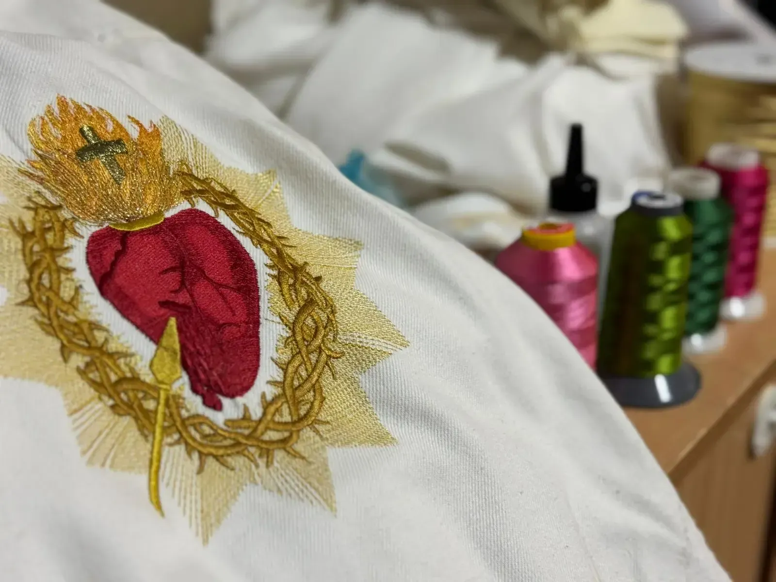 Sagrado Corazón bordado sobre tela blanca con carretes de hilo dorado, rojo y verde de fondo.