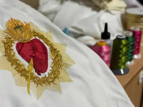 Sagrado Corazón bordado sobre tela blanca con carretes de hilo dorado, rojo y verde de fondo.
