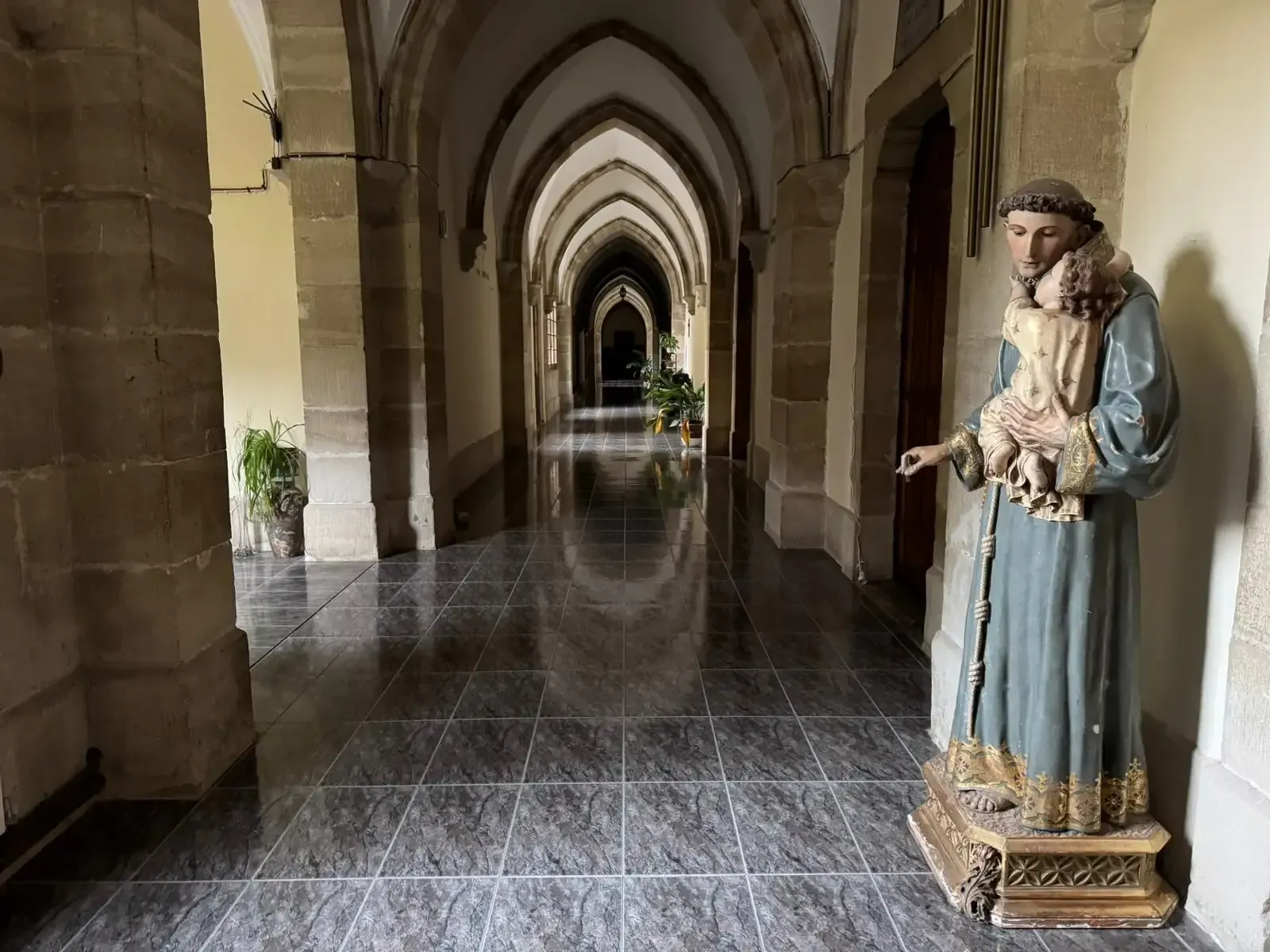 Pasillo con techo abovedado y suelo de baldosas; estatua de una figura vestida con una túnica que sostiene un cordero.