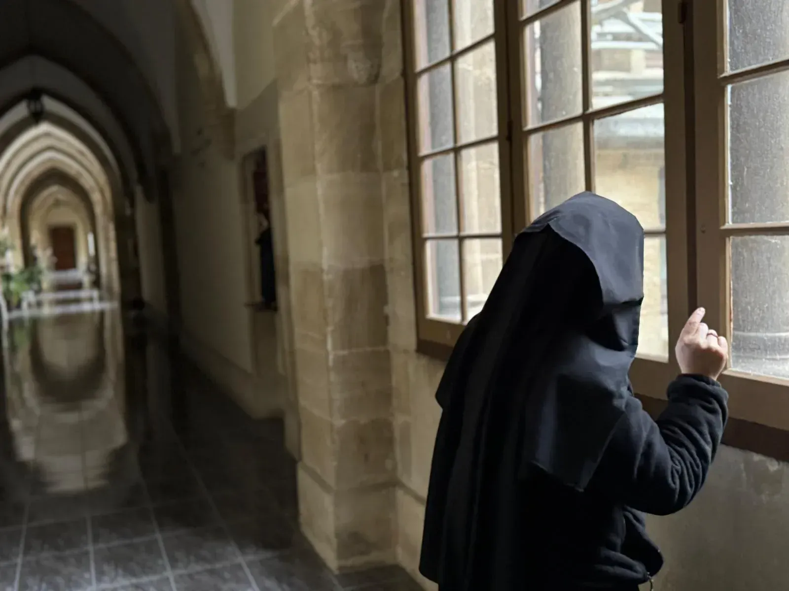 Monja con hábito negro, mirando por una ventana. Pasillo arqueado al fondo.