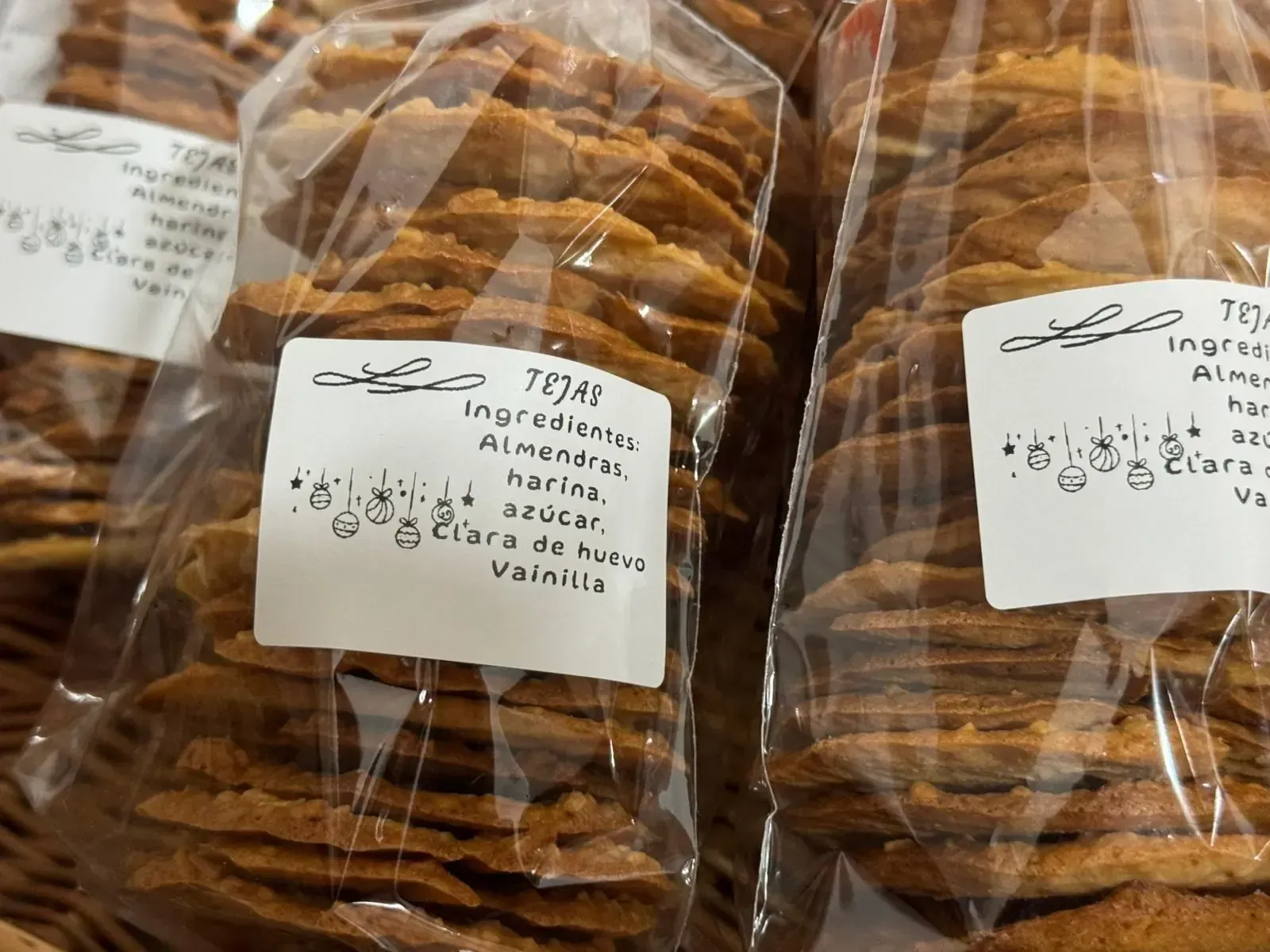 Paquetes de galletas de almendras finas y redondas en plástico transparente, etiquetas con ingredientes: almendras, harina, azúcar, clara de huevo, vainilla.