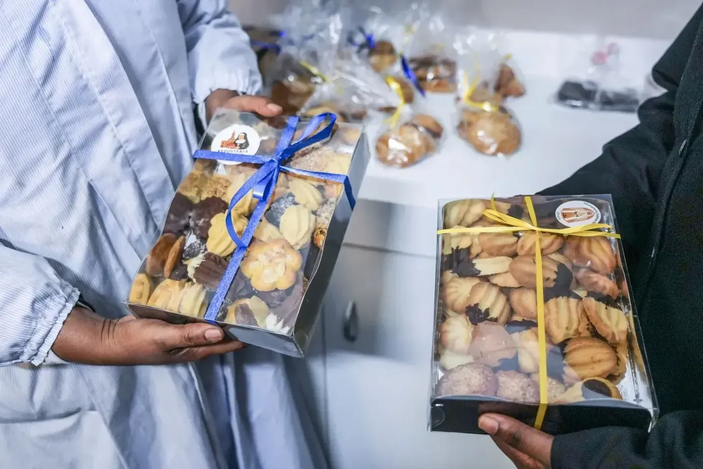 Dos personas sosteniendo cajas de regalo de galletas, una con cinta azul y la otra con cinta amarilla.