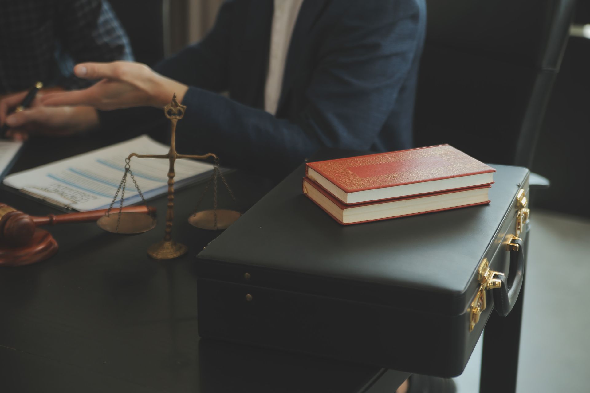 Escena de despacho de abogados: balanza, mazo, maletín y libro sobre una mesa, dos personas hablando de fondo.