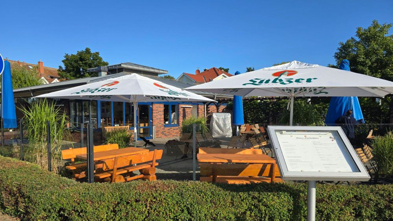 Fischerhütte Rerik mit weißen Sonnenschirmen, Holzbänken und einem Speisekartenschild an einem sonnigen Tag.