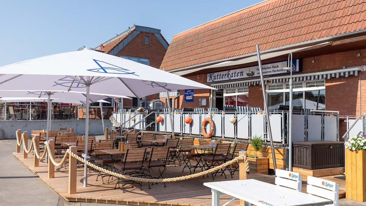 Terrasse vom Kutterkaten im Freien mit Holzstühlen, weißen Sonnenschirmen und einem rotziegelgedeckten Gebäude im Hintergrund