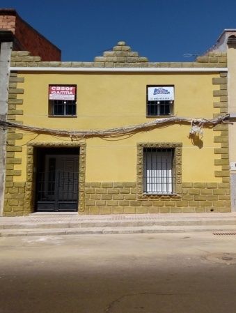 Edificio amarillo con dos ventanas y una puerta; acentos de ladrillo en la fachada.