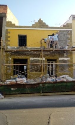 Fachada de edificio en renovación; paredes amarillas, detalles de piedra, andamios, persona trabajando, escombros en la base.