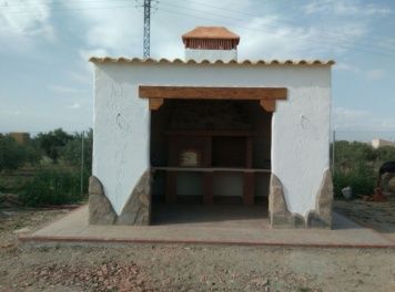Horno exterior de paredes blancas con molduras de madera y chimenea, situado sobre una losa de hormigón en un campo.