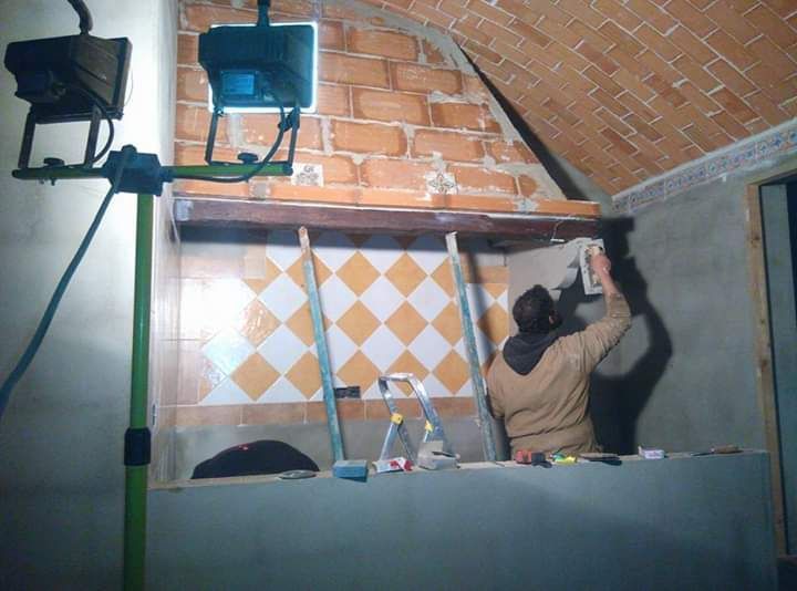 Persona aplicando yeso a una pared con una llana, bajo luces de trabajo, cerca de un nicho de azulejos con techo de ladrillo.