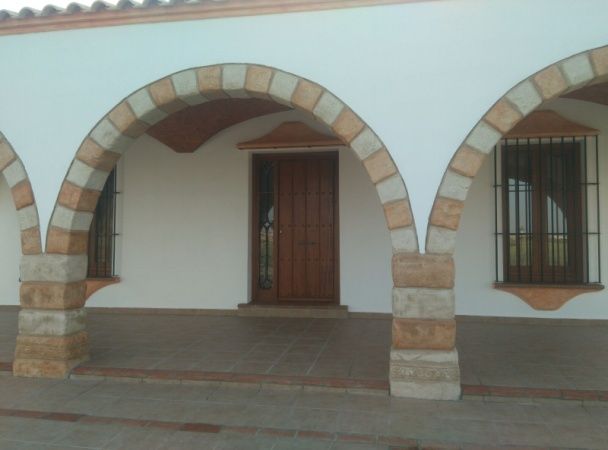 Porche arqueado con columnas y molduras de piedra, puerta marrón, dos ventanas enrejadas y paredes blancas.