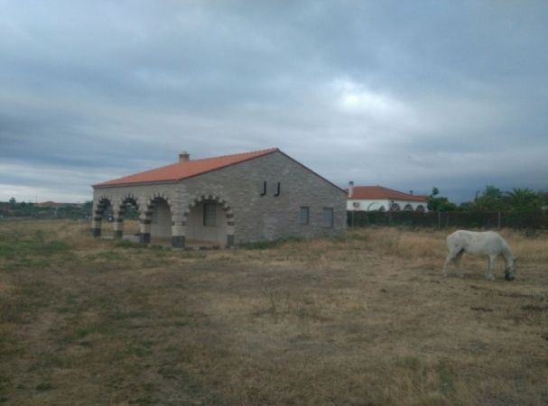 Edificio de piedra con columnas arqueadas y tejado de tejas rojas. Caballo blanco pastando en el campo. Cielo nublado.