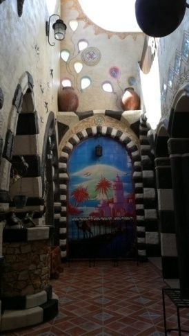 Interior con paredes coloridas, arcos decorativos y un mural pintado de un castillo junto al mar.