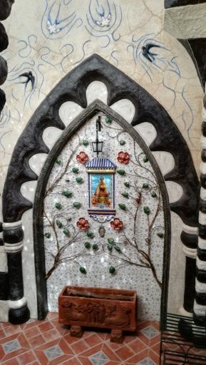 Mural de mosaico con marco arqueado y motivos florales y de aves. Estatua en farol colgante y macetero.