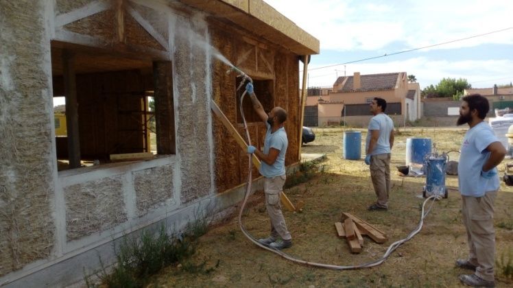 Trabajadores rocían aislante sobre la estructura de una casa. Uno rocía, dos vigilan, camisas azules, al aire libre.