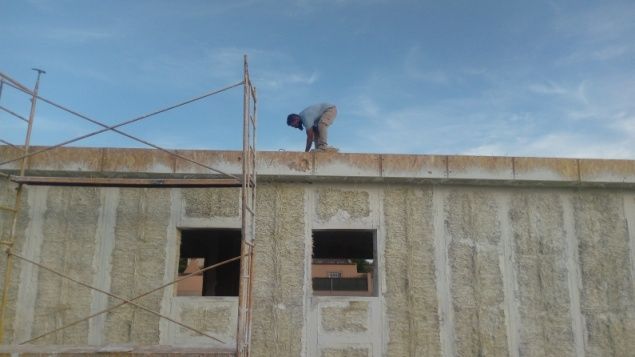 Obrero en el tejado de un edificio, cerca de un andamio. Cielo azul. Pared beige con dos ventanas.