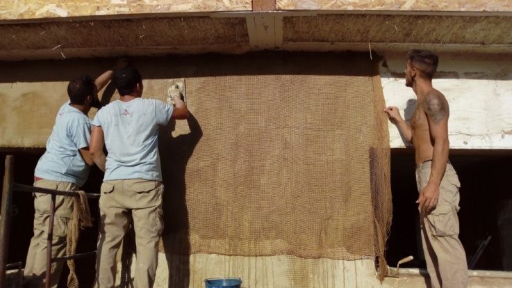 Tres personas aplicando material a una pared al aire libre. Una sin camisa, dos con camisa. Proyecto de construcción.