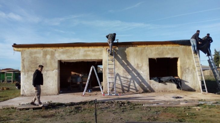 Hombres trabajando en el tejado de un edificio, de pie sobre escaleras. El edificio tiene garaje y ventanas abiertas, y el cielo es azul.