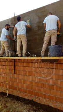 Tres trabajadores enyesan una pared de ladrillos; llevan camisas azul claro y pantalones color canela.