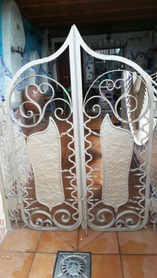 Puertas de hierro forjado ornamentales de color blanco, con parte superior arqueada, con diseños de escudos y detalles de volutas.