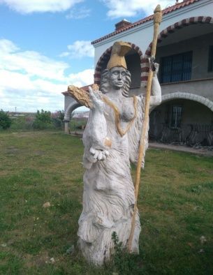 Estatua de una mujer con túnicas vaporosas y tocado, sosteniendo un bastón, en un patio cubierto de hierba.