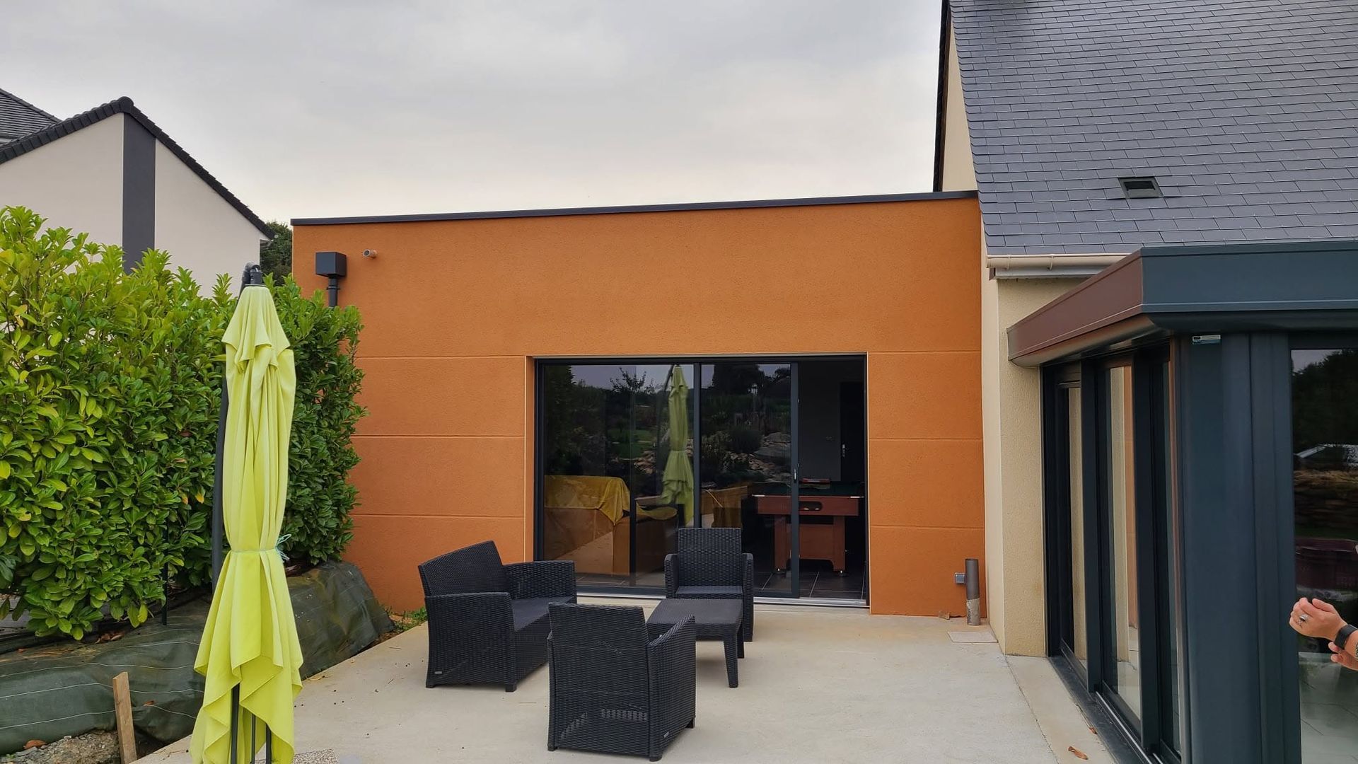 Terrasse extérieure avec chaises en osier noir, parasol vert citron et mur orange avec portes vitrées.