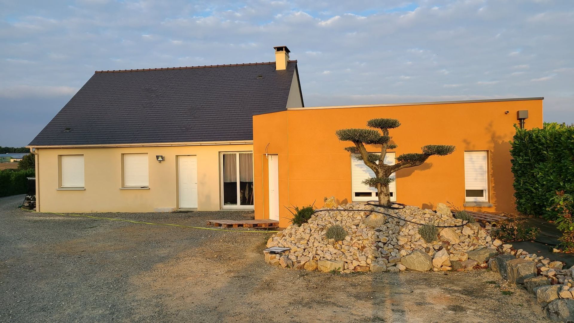 Maison de plain-pied aux murs orange et beige, toit sombre et jardin de rocaille à l'avant sous un ciel dégagé.