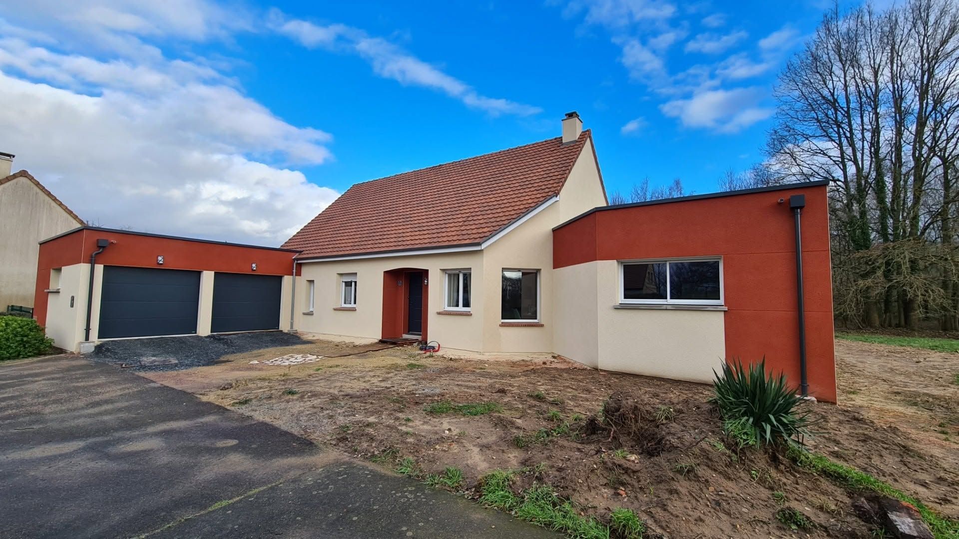 Maison de plain-pied couleur crème avec toit rouge, garage attenant et jardin nu sous un ciel bleu.