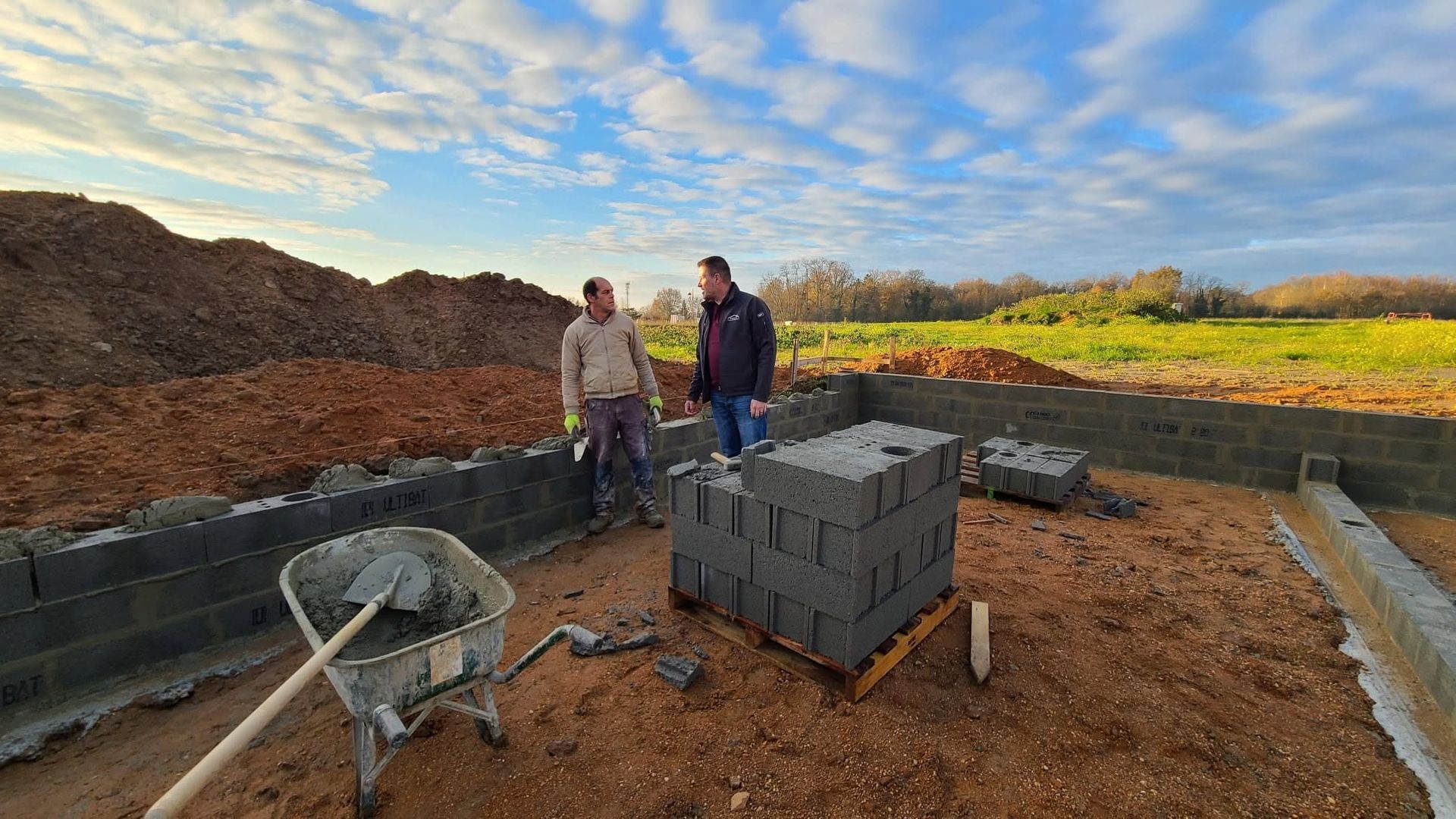 Deux ouvriers se tiennent debout sur un chantier de construction, près de fondations inachevées, avec des parpaings empilés et une brouette.