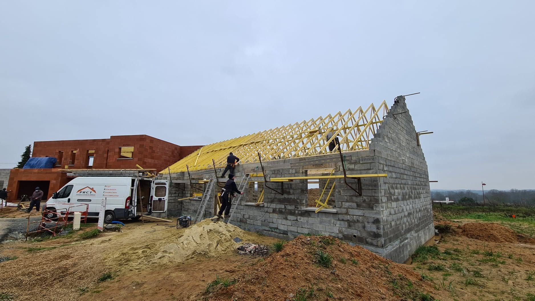 Maison en pierre en construction, charpente de toit en place, ouvriers et camionnette blanche sur un site rural.