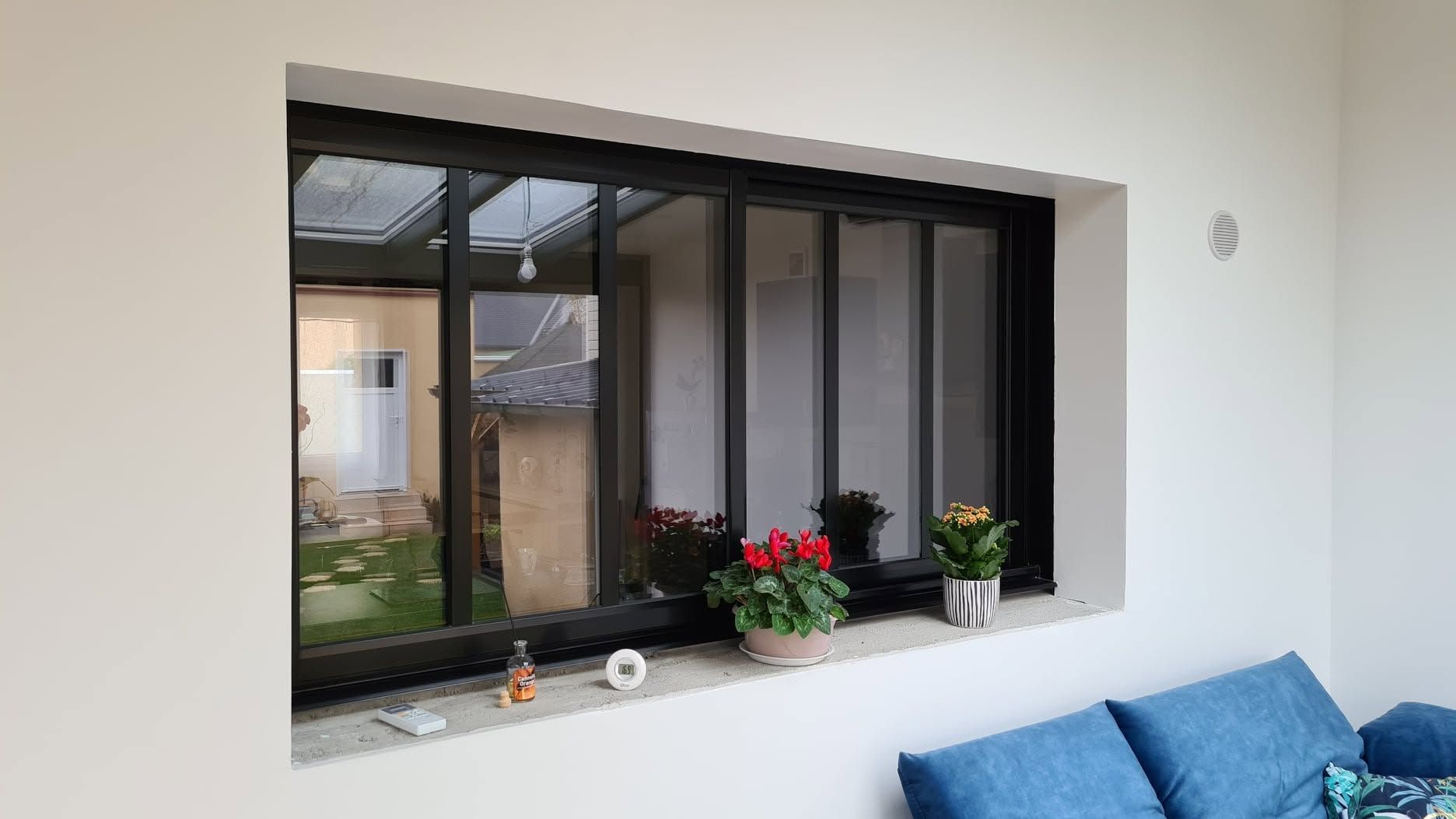 Intérieur moderne avec une fenêtre à cadre noir, des fleurs en pot sur le rebord et un canapé bleu à droite.