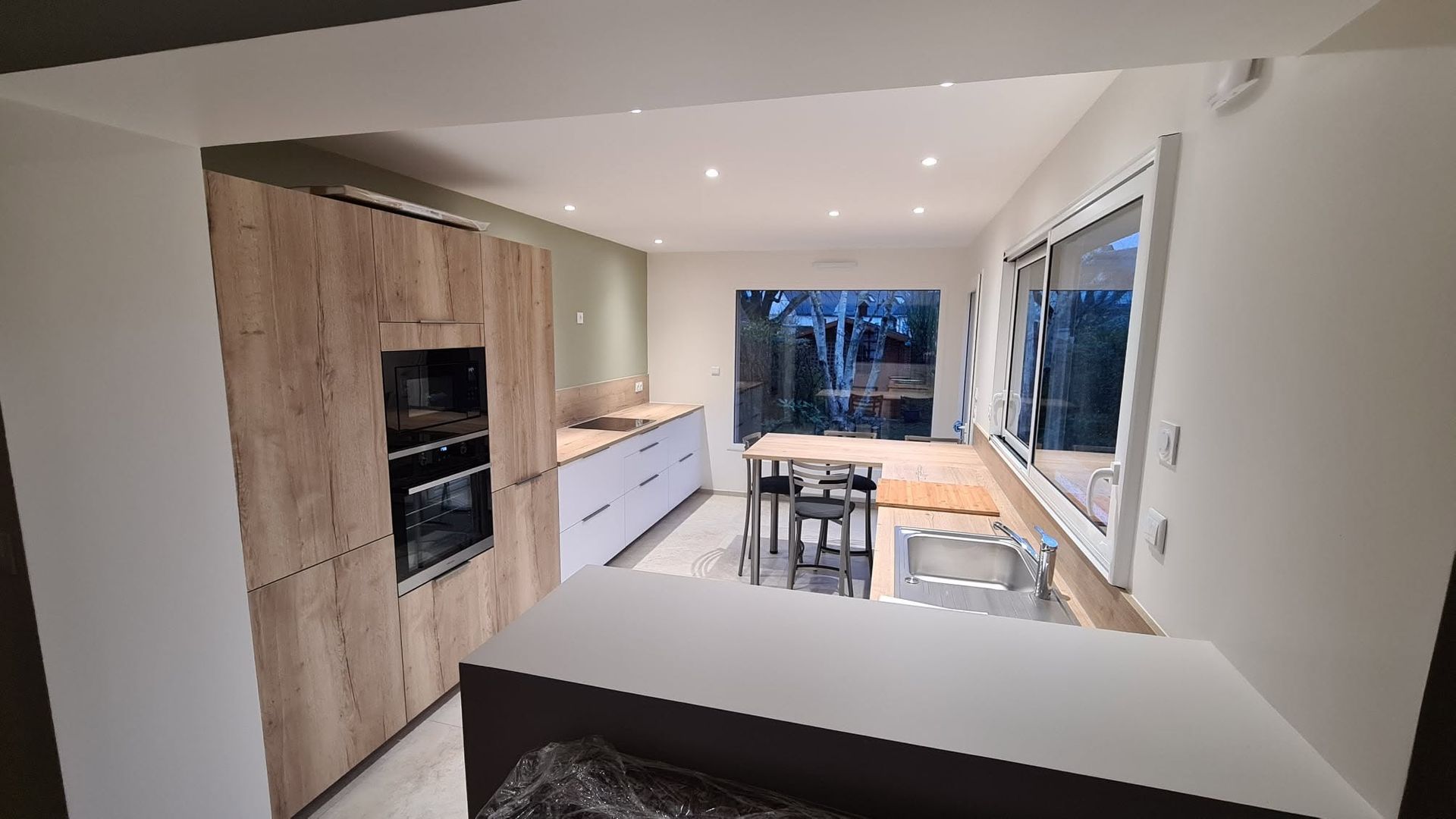 Cuisine moderne avec armoires en bois, comptoirs blancs, îlot et grande fenêtre donnant sur les arbres.