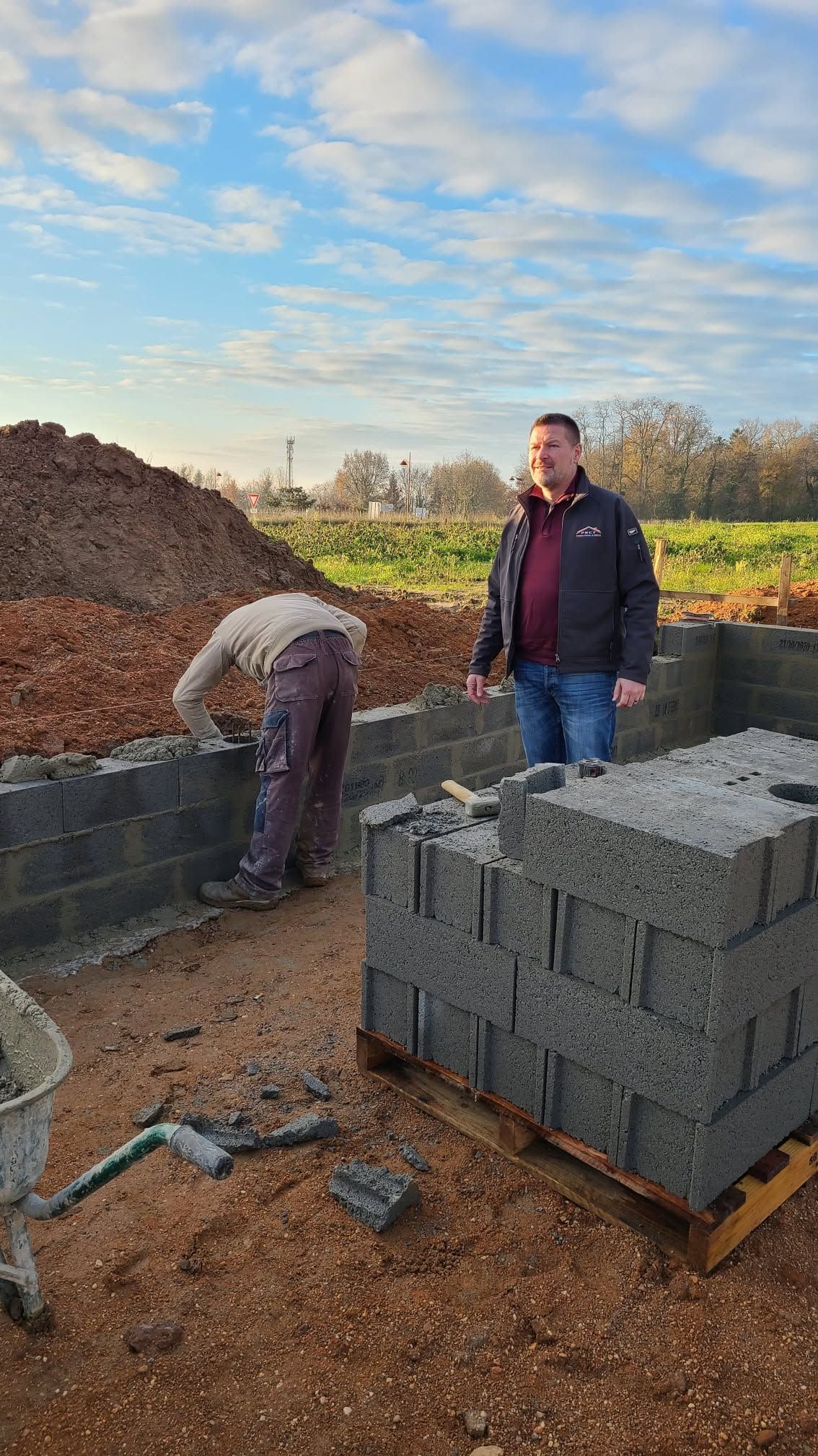 Deux ouvriers sur un chantier de construction, à côté de blocs de béton gris empilés sous un ciel nuageux.