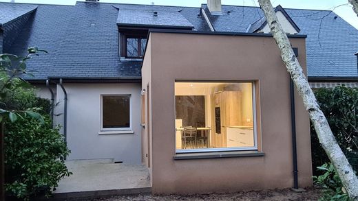 Arrière d'une maison avec une grande fenêtre éclairée et un tronc d'arbre tombé appuyé contre le mur.