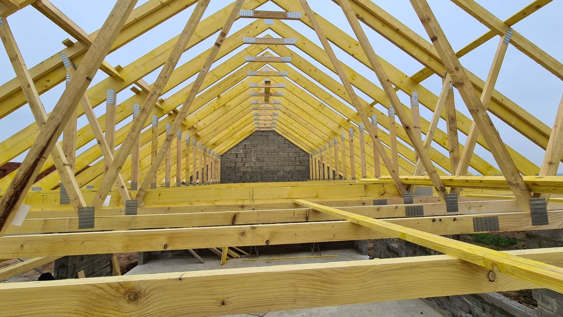 Intérieur d'une charpente en bois en construction, avec des fermes en bois jaunes et une structure ouverte éclairée par un puits de lumière.