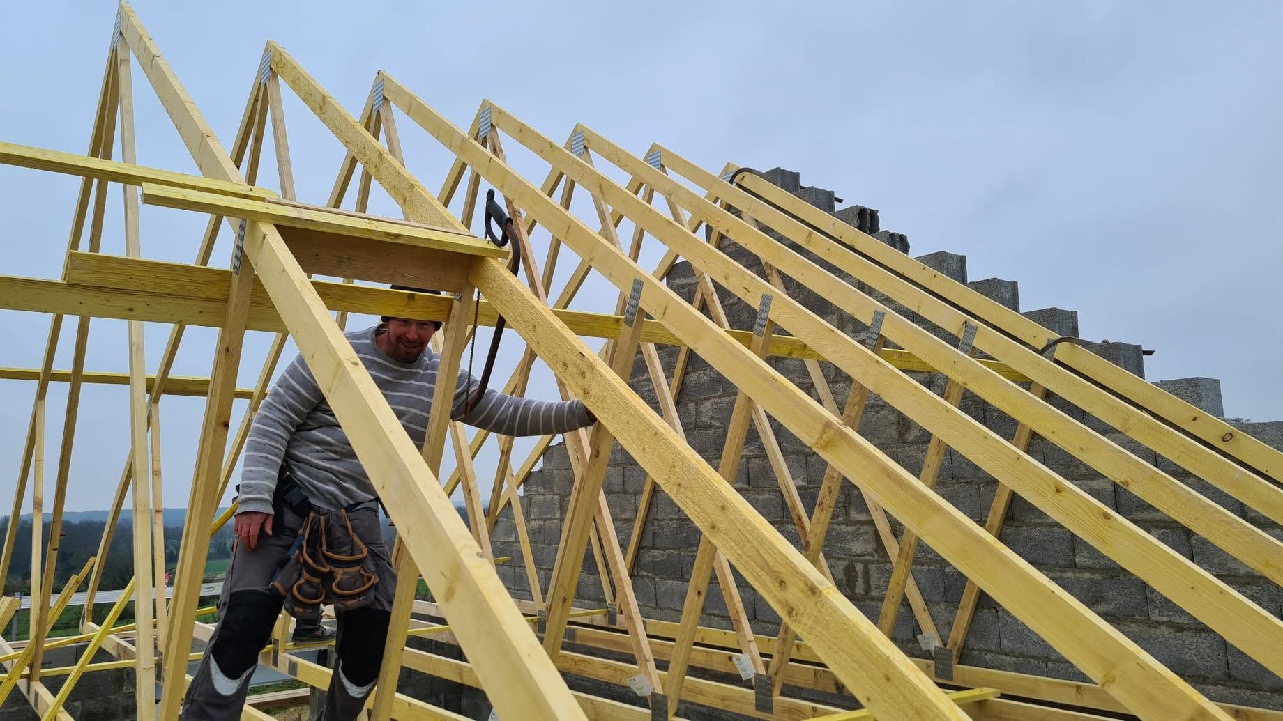 Ouvrier travaillant sur une charpente de toit en bois en construction, sous un ciel nuageux.