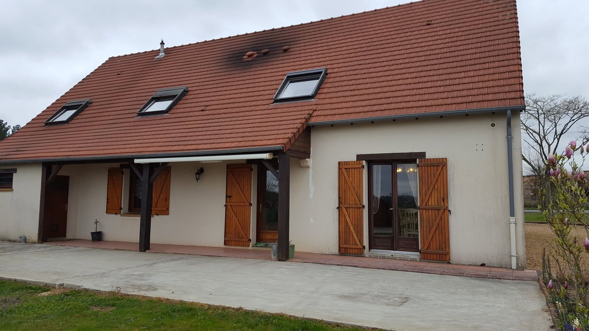 Maison blanche avec un toit de tuiles rouges, des lucarnes et des volets en bois sur un terrain gazonné.